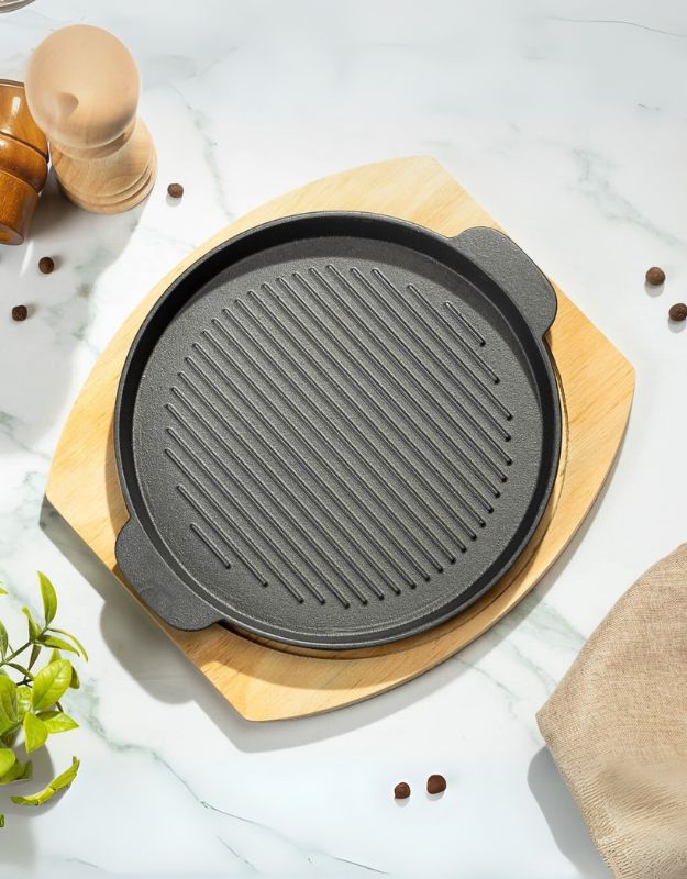 A round, ribbed cast iron griddle pan on a wooden base, with a bamboo stand and various cooking utensils in the background.